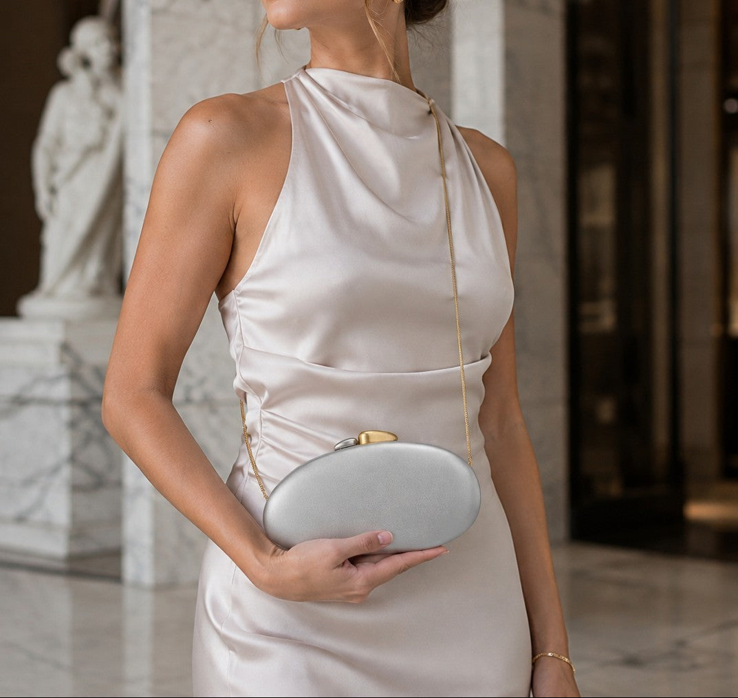 VALENORA Cosette clutch bag in silver vegan leather with a two-tone gold and silver clasp held by a woman in a dress.