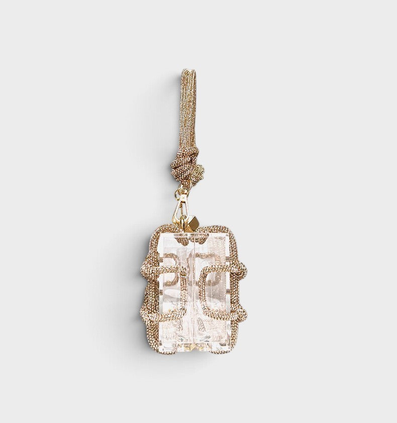 VALENORA Isabelle transparent acrylic clutch bag with gold chain detailing and a knotted wrist strap.