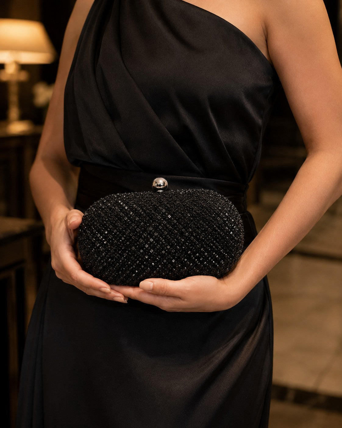 VALENORA Ismay oval clutch bag covered in black crystals with a silver ball clasp held by a woman in a black dress.