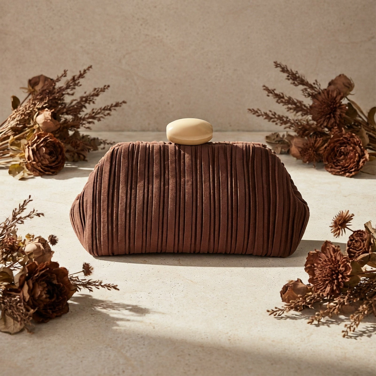 VALENORA Liora brown pleated suede clutch bag with a smooth oval stone clasp, displayed on a textured stone surface.
