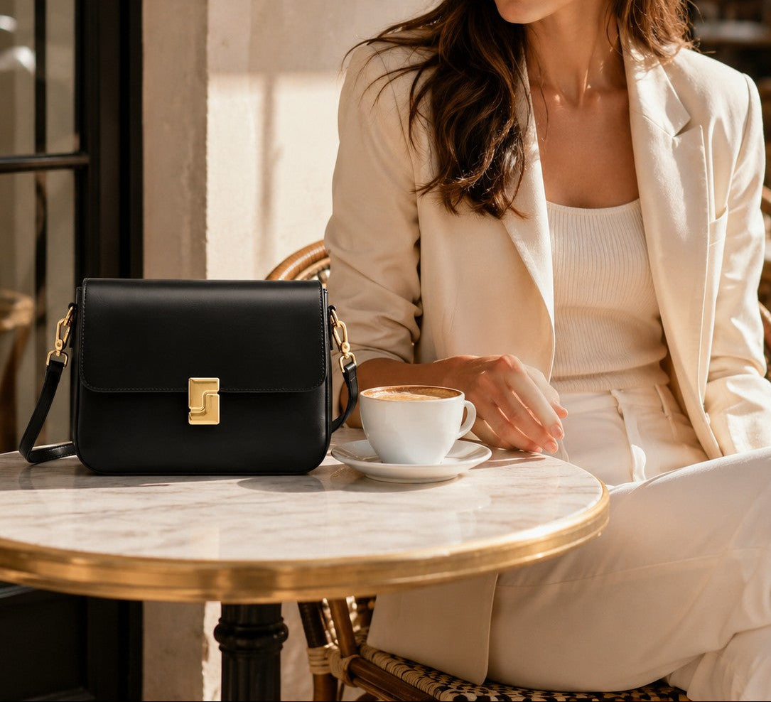 VALENORA Marisa black leather shoulder bag with gold buckle on a cafe table next to a woman in a beige suit