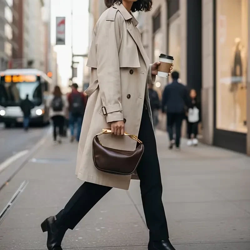 VALENORA Roxane handbag in dark brown leather with a gold twisted handle, carried by a woman in a beige trench coat.